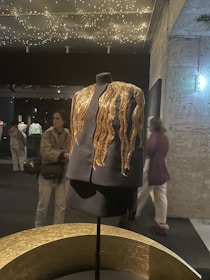A black mannequin dressed in an elegant black outfit features striking golden embellishments that resemble flowing waves. These details catch the light from the star-like ceiling decor in a dimly lit exhibition space with textured walls and various people in the background.