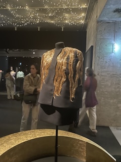 A black mannequin dressed in an elegant black outfit features striking golden embellishments that resemble flowing waves. These details catch the light from the star-like ceiling decor in a dimly lit exhibition space with textured walls and various people in the background.