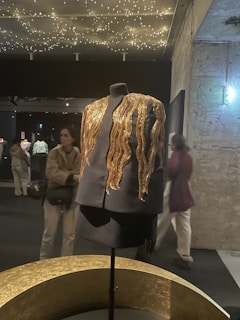 A black mannequin dressed in an elegant black outfit features striking golden embellishments that resemble flowing waves. These details catch the light from the star-like ceiling decor in a dimly lit exhibition space with textured walls and various people in the background.