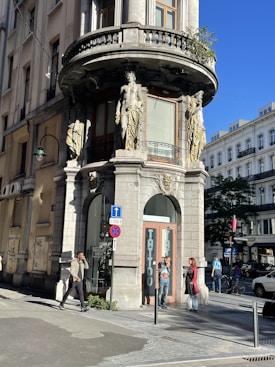 A street corner featuring a historic building with ornate classical statues on its façade. The building has large windows and a sign that reads 'Tattoo'. Several people are walking or standing on the sidewalk, which is adorned with various street signs.