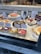 A bakery display featuring a variety of cakes and tarts arranged neatly on a marble countertop. The desserts include fruit tarts, cheesecakes, and other pastries, each labeled with a small sign indicating its type or flavor. Bottled beverages are placed in the background.