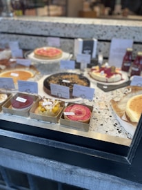 A bakery display featuring a variety of cakes and tarts arranged neatly on a marble countertop. The desserts include fruit tarts, cheesecakes, and other pastries, each labeled with a small sign indicating its type or flavor. Bottled beverages are placed in the background.
