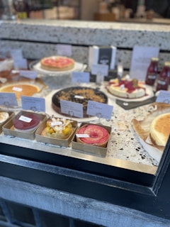 A bakery display featuring a variety of cakes and tarts arranged neatly on a marble countertop. The desserts include fruit tarts, cheesecakes, and other pastries, each labeled with a small sign indicating its type or flavor. Bottled beverages are placed in the background.