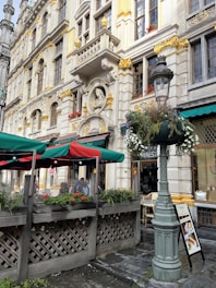 A detailed facade of a historic building adorned with ornate carvings and decorative elements, featuring a bust set in an alcove. The building has large windows with small flower boxes beneath them. The street level reveals a quaint outdoor café with green and red umbrellas covering the seating area, surrounded by wooden planter boxes filled with greenery and vibrant flowers. A classic streetlamp is adorned with hanging plants, and a menu board is visible near the entrance.