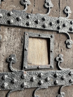 A close-up view of a wooden surface reinforced by ornate, dark metal bands studded with rivets. The design includes decorative clover-shaped elements and a square frame in the middle, showcasing the weathered texture of the wood beneath.