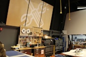 A workshop or maker space containing tools and equipment is shown. Pegboards with various hand tools like screwdrivers, wrenches, and tape are mounted on the wall. Large tool chests and cabinets are placed beneath the pegboards. The room has a high ceiling with industrial black piping and hanging yellow cords. A logo with a wrench and screwdriver symbol is prominently displayed on a large wooden board.