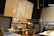 A workshop or maker space containing tools and equipment is shown. Pegboards with various hand tools like screwdrivers, wrenches, and tape are mounted on the wall. Large tool chests and cabinets are placed beneath the pegboards. The room has a high ceiling with industrial black piping and hanging yellow cords. A logo with a wrench and screwdriver symbol is prominently displayed on a large wooden board.