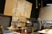 A workshop or maker space containing tools and equipment is shown. Pegboards with various hand tools like screwdrivers, wrenches, and tape are mounted on the wall. Large tool chests and cabinets are placed beneath the pegboards. The room has a high ceiling with industrial black piping and hanging yellow cords. A logo with a wrench and screwdriver symbol is prominently displayed on a large wooden board.