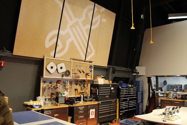 A workshop or maker space containing tools and equipment is shown. Pegboards with various hand tools like screwdrivers, wrenches, and tape are mounted on the wall. Large tool chests and cabinets are placed beneath the pegboards. The room has a high ceiling with industrial black piping and hanging yellow cords. A logo with a wrench and screwdriver symbol is prominently displayed on a large wooden board.