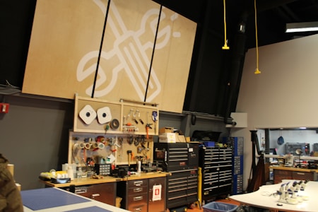 A workshop or maker space containing tools and equipment is shown. Pegboards with various hand tools like screwdrivers, wrenches, and tape are mounted on the wall. Large tool chests and cabinets are placed beneath the pegboards. The room has a high ceiling with industrial black piping and hanging yellow cords. A logo with a wrench and screwdriver symbol is prominently displayed on a large wooden board.