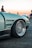 A close-up of a sleek, modified car with a focus on the front wheel and intricately designed rim. The car has a matte finish with a modern, aerodynamic body. In the background, a few people are visible, with one person standing closer to the car. The scene is set outdoors, possibly at a car meet or event, with another car visible in the distance.