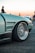 A close-up of a sleek, modified car with a focus on the front wheel and intricately designed rim. The car has a matte finish with a modern, aerodynamic body. In the background, a few people are visible, with one person standing closer to the car. The scene is set outdoors, possibly at a car meet or event, with another car visible in the distance.