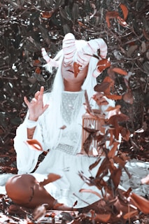 A person with long white hair and large, striped horns sits in a forest-like setting surrounded by fallen leaves. They wear a flowing, light-colored garment and hold a small wooden cage, emanating a mystical and surreal ambiance.