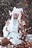 A person with long white hair and large, striped horns sits in a forest-like setting surrounded by fallen leaves. They wear a flowing, light-colored garment and hold a small wooden cage, emanating a mystical and surreal ambiance.