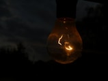 A glowing light bulb is suspended in a dark environment, casting a warm and subtle light. The background is dimly lit, with blurred outlines of trees or foliage suggesting an outdoor setting during dusk or nighttime.