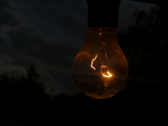 A glowing light bulb is suspended in a dark environment, casting a warm and subtle light. The background is dimly lit, with blurred outlines of trees or foliage suggesting an outdoor setting during dusk or nighttime.
