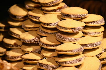 A large number of sandwich cookies with golden-yellow tops are stacked in a pyramid-like structure. The cookies have a dark filling and some are coated with shredded coconut around the edges.