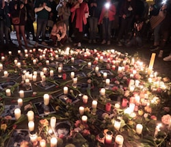 A gathering of people stands around a memorial arrangement featuring numerous lit candles and flowers placed on the ground. Photographs are interspersed among the candles and flowers, creating a poignant scene. The ambient lighting and presence of individuals suggest a respectful and somber atmosphere.