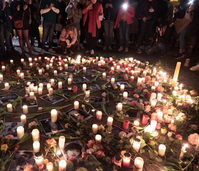 A gathering of people stands around a memorial arrangement featuring numerous lit candles and flowers placed on the ground. Photographs are interspersed among the candles and flowers, creating a poignant scene. The ambient lighting and presence of individuals suggest a respectful and somber atmosphere.
