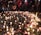 A gathering of people stands around a memorial arrangement featuring numerous lit candles and flowers placed on the ground. Photographs are interspersed among the candles and flowers, creating a poignant scene. The ambient lighting and presence of individuals suggest a respectful and somber atmosphere.