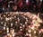 A gathering of people stands around a memorial arrangement featuring numerous lit candles and flowers placed on the ground. Photographs are interspersed among the candles and flowers, creating a poignant scene. The ambient lighting and presence of individuals suggest a respectful and somber atmosphere.