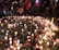 A gathering of people stands around a memorial arrangement featuring numerous lit candles and flowers placed on the ground. Photographs are interspersed among the candles and flowers, creating a poignant scene. The ambient lighting and presence of individuals suggest a respectful and somber atmosphere.