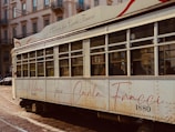 A vintage tram with a cream exterior and red accents is captured on a city street. The tram features an elegant script with the words 'Milano per Carla Fracci' and the number '1880'. Buildings with ornate architecture and balconies line the street in the background.