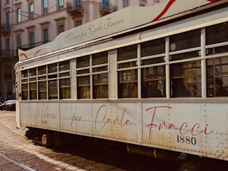 A vintage tram with a cream exterior and red accents is captured on a city street. The tram features an elegant script with the words 'Milano per Carla Fracci' and the number '1880'. Buildings with ornate architecture and balconies line the street in the background.