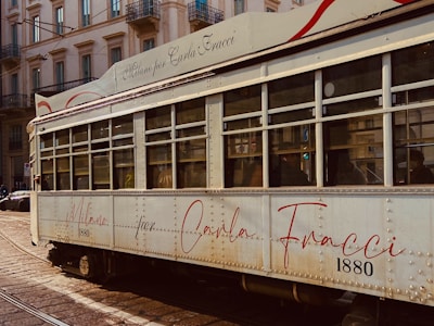 A vintage tram with a cream exterior and red accents is captured on a city street. The tram features an elegant script with the words 'Milano per Carla Fracci' and the number '1880'. Buildings with ornate architecture and balconies line the street in the background.