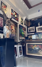 A richly decorated art gallery interior filled with a variety of framed paintings covering the walls. In the foreground, there is a statue of a figure with wings sitting on a rock. The paintings display diverse styles, including abstract and figurative art. The room is well-lit with spotlights illuminating the artwork.