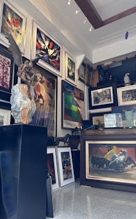 A richly decorated art gallery interior filled with a variety of framed paintings covering the walls. In the foreground, there is a statue of a figure with wings sitting on a rock. The paintings display diverse styles, including abstract and figurative art. The room is well-lit with spotlights illuminating the artwork.