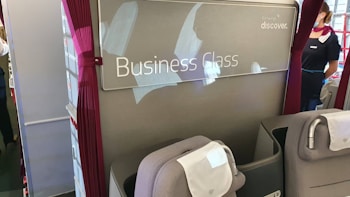 The image depicts the interior of an aircraft business class section, featuring plush reclining seats with headrests and attached cloth covers. To the right, a flight attendant stands wearing a uniform and mask near the entrance. The area is designated as 'Business Class' with signage, and the ambiance is professional and neat, characterized by soft lighting.
