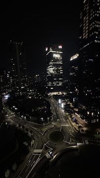 A nighttime cityscape featuring tightly clustered high-rise buildings. The center of the image highlights a tall building with 'FWD' and 'Citi' logos prominently displayed. Below, a major roundabout with lighted streets and some light trails from vehicles is visible. Lighted windows and streetlights give subtle illumination to the surroundings.
