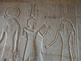 Carved into a stone wall, three ancient Egyptian figures are depicted in profile, wearing intricate headdresses and garments. Hieroglyphics are visible around the figures.
