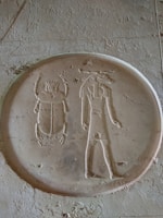An ancient Egyptian hieroglyphic carving depicting a scarab beetle and a standing human figure with a crown or headdress, in a rounded frame. The carving is etched into a flat surface with weathered textures.