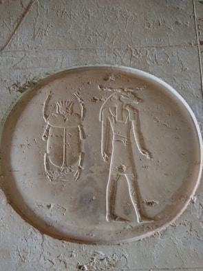 An ancient Egyptian hieroglyphic carving depicting a scarab beetle and a standing human figure with a crown or headdress, in a rounded frame. The carving is etched into a flat surface with weathered textures.