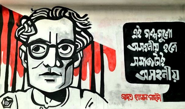 A mural featuring a stylized black and white portrait of a person with glasses and wavy hair. The background contains dramatic red paint strokes resembling dripping blood. On the right side, there is bold black text in a non-Latin script.