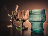 A set of three elegant glass objects is positioned on a wooden surface against a warm, softly lit background. From left to right, there is a clear, tall glass goblet with a decorative stem. The middle item is a green-tinted glass bowl with a scalloped edge, and on the right, there is a textured blue glass vessel with a unique shape.
