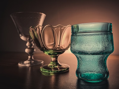 A set of three elegant glass objects is positioned on a wooden surface against a warm, softly lit background. From left to right, there is a clear, tall glass goblet with a decorative stem. The middle item is a green-tinted glass bowl with a scalloped edge, and on the right, there is a textured blue glass vessel with a unique shape.