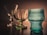 A set of three elegant glass objects is positioned on a wooden surface against a warm, softly lit background. From left to right, there is a clear, tall glass goblet with a decorative stem. The middle item is a green-tinted glass bowl with a scalloped edge, and on the right, there is a textured blue glass vessel with a unique shape.