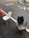 Two old chairs are placed on an asphalt road near an orange traffic cone. The chair on the left is a white, worn dining chair, while the one on the right is a black office chair with a cracked seat. The pavement markings include the numbers 30 and an X. In the background, there are more traffic cones and a couple of office chairs scattered along the road.