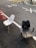 Two old chairs are placed on an asphalt road near an orange traffic cone. The chair on the left is a white, worn dining chair, while the one on the right is a black office chair with a cracked seat. The pavement markings include the numbers 30 and an X. In the background, there are more traffic cones and a couple of office chairs scattered along the road.