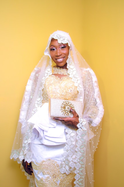 A person dressed in elegant traditional attire, featuring a detailed white and gold gown with intricate lace and bead embellishments. The individual is wearing a matching headpiece and holding an embellished clutch. Henna designs adorn their hands.