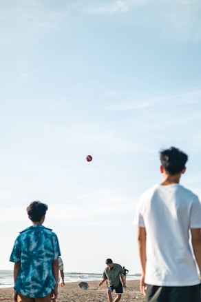 A group of young people is playing volleyball on a beach. The volleyball is captured in mid-air, and the players are actively engaged in the game. The atmosphere is casual and relaxed, with clear blue skies and the ocean visible in the background.