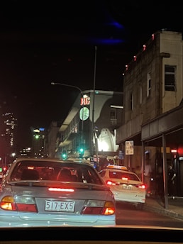 A nighttime urban scene with several cars stopped in traffic on a city street. A prominent building displays a large inflatable ghost decoration on its facade. Neon lights illuminate the street, highlighting the hustle and bustle typical of a city environment. The signage and license plates indicate the location might be in Queensland, Australia.