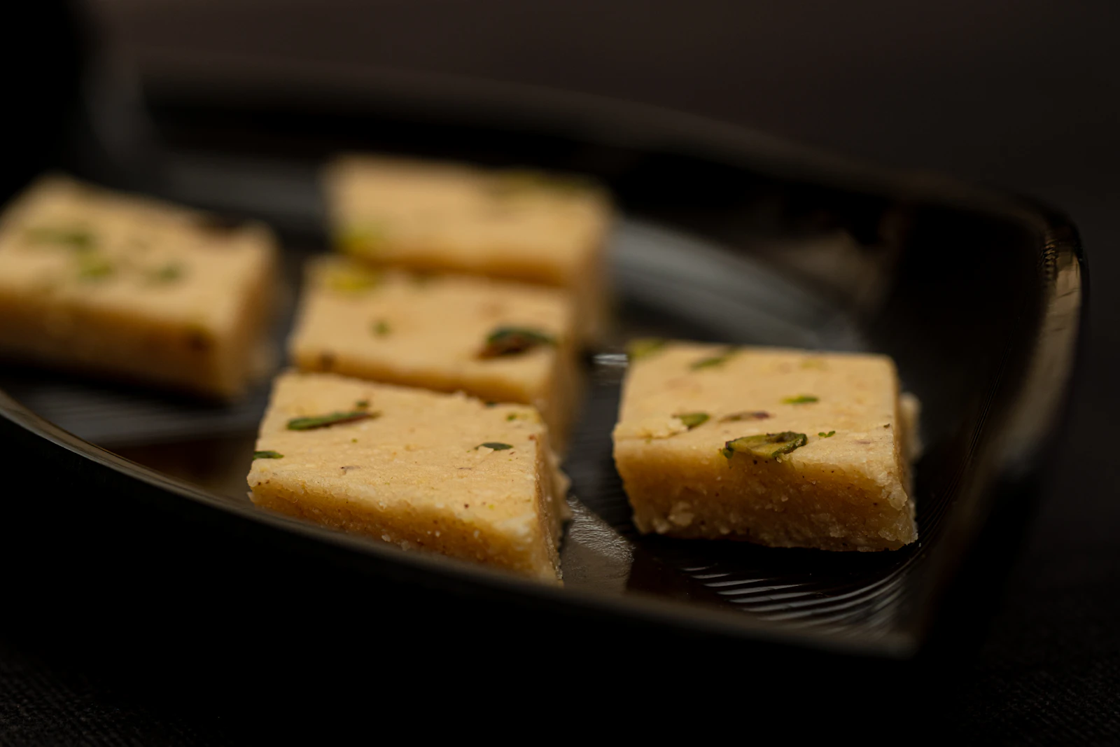 Squares of burfi flecked with pistachio on a dark plate