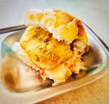 A delicious snack featuring a fried potato patty placed inside a soft bun, served on a stainless steel plate. The snack is complemented by a sprinkle of red spice, which adds to its visual and flavorful appeal.