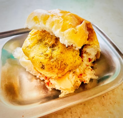 A delicious snack featuring a fried potato patty placed inside a soft bun, served on a stainless steel plate. The snack is complemented by a sprinkle of red spice, which adds to its visual and flavorful appeal.