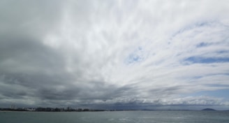 Thick clouds cover the sky, creating an overcast atmosphere. The horizon separates the sky from the sea below, with distant buildings faintly visible along the coastline. Patches of white and gray are predominant in the cloud formations, with hints of blue sky peeking through.