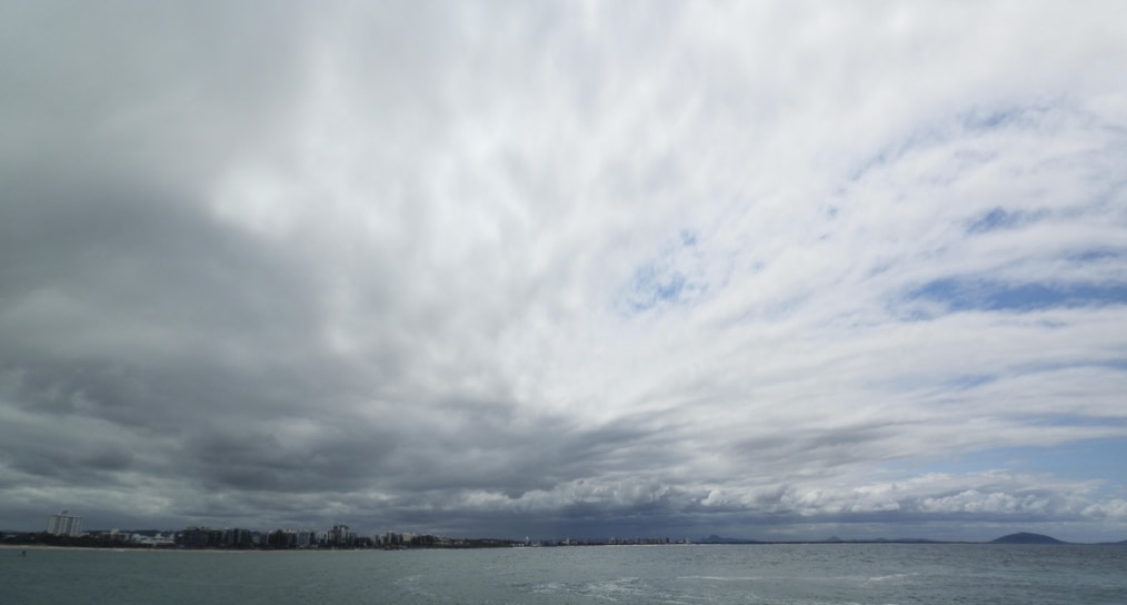 Thick clouds cover the sky, creating an overcast atmosphere. The horizon separates the sky from the sea below, with distant buildings faintly visible along the coastline. Patches of white and gray are predominant in the cloud formations, with hints of blue sky peeking through.